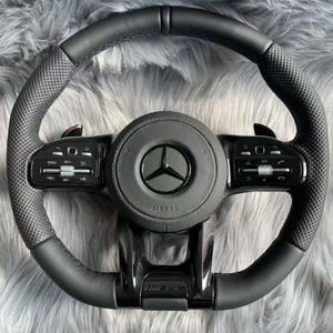 May include: A black leather steering wheel featuring a perforated grip texture, a central circular hub with the Mercedes-Benz logo, and integrated control panels on the horizontal spokes. The steering wheel includes paddle shifters and an AMG emblem.