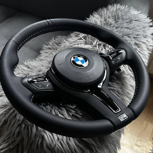 May include: Black leather steering wheel with blue stitching and a BMW logo. The wheel has integrated controls and a sporty design. The image shows a close-up of the steering wheel, highlighting its details and craftsmanship.