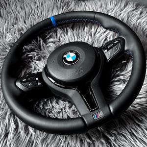 May include: Black leather steering wheel with blue stitching and a BMW logo. The wheel has integrated controls and a sporty design. The M logo is visible on the lower spoke.