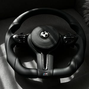 May include: Black leather steering wheel with perforated grips and integrated controls. The center features the BMW logo and the word "AIRBAG". The bottom has the "M" logo.
