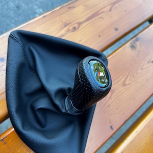 Op de afbeelding: Zwarte lederen versnellingspookknop met het BMW M-logo. De knop heeft een geperforeerde grip en een duidelijke schakelpatroonindicator. De versnellingspook is geplaatst op een zwarte leren hoes.