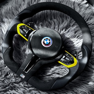 May include: A black and yellow BMW steering wheel with a perforated grip. The wheel features the BMW logo and control buttons. The airbag cover is black with the word "AIRBAG" printed on it.