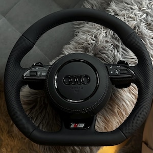 以下が含まれることがあります： パンチングレザーのグリップ、AudiロゴとAIRBAGの文字が入った中央の円形ホーンパッド、水平スポークに統合されたコントロールボタン、そしてステアリングホイール下部にRSバッジを備えた黒いAudiステアリングホイール。