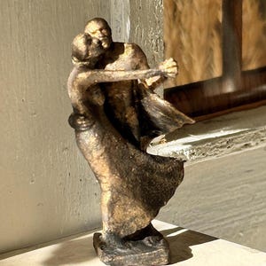 May include: Bronze sculpture depicting a dancing couple in a close embrace. The figures are rendered with detail, their clothing flowing, creating a sense of movement. The sculpture is mounted on a square base, suitable for display.