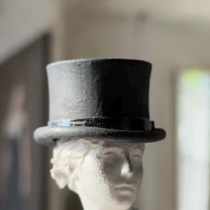 May include: A miniature black top hat sits atop a white bust of a classical figure. The hat has a shiny band and a textured surface. The bust is detailed with facial features and hair. The background is blurred, with natural light.