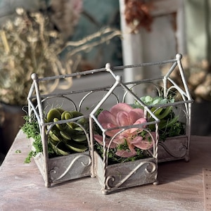 Pode incluir: Três floreiras decorativas de metal com acabamento envelhecido, cada uma em forma de casa pequena. Contêm suculentas artificiais em tons de verde e rosa, aninhadas em musgo verde. As floreiras estão sobre uma superfície de madeira clara.