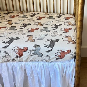 May include: A crib sheet with a repeating pattern of running horses in shades of brown, gray, and blue on a cream background. The crib sheet is paired with a white ruffled crib skirt, creating a soft, inviting look.