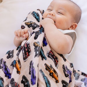 May include: A baby swaddled in a beige muslin swaddle blanket with a print of vintage cars in various colors. The baby is smiling and has their hand in their mouth.