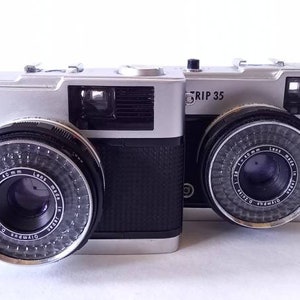 Op de afbeelding: Twee zilveren en zwarte Olympus Trip 35 filmcamera's met een zwarte leren riem bevestigd aan een van de camera's.