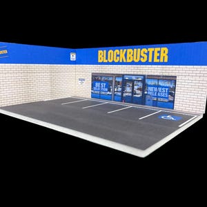 May include: A miniature model of a Blockbuster video store with a brick facade, blue and yellow signage, and a small parking lot with a handicapped parking space.