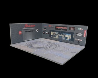 Può includere: Un diorama di garage modello con un muro di mattoni grigi, un portone del garage nero con il testo "#BAGGED" e un pavimento in cemento grigio con segni di pneumatici. Il muro presenta vari loghi e testi, tra cui "SingleLap", "Performance", "No Quema Cunt" e "Nitro Lift".