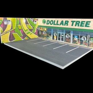 May include: A miniature model of a Dollar Tree store with a green sign and a parking lot. The store has a brick facade and large windows. The parking lot is made of gray asphalt with white lines.