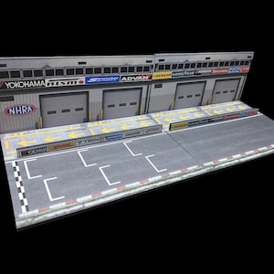 May include: A model of a race car garage with two bays, each with a roll-up door. The garage is decorated with logos of various racing sponsors, including Yokohama, Nismo, Spoon, and Advan. The garage is set on a gray asphalt track with white lines and yellow arrows.