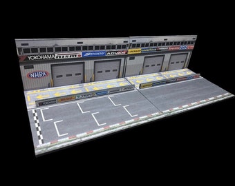May include: A model of a race car garage with two bays, each with a roll-up door. The garage is decorated with logos of various racing sponsors, including Yokohama, Nismo, Spoon, and Advan. The garage is set on a gray asphalt track with white lines and yellow arrows.