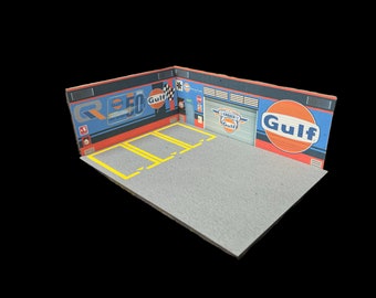 May include: A miniature diorama of a Gulf racing garage. The scene features a gray concrete floor with yellow lane markings, blue walls, and Gulf branding. The garage door is closed, and the scene is set against a black background.