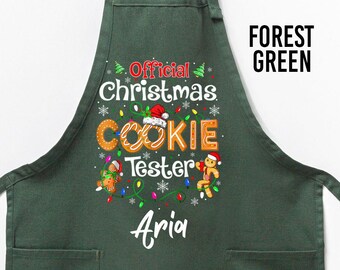 Op de afbeelding: Bosse groene schort met de tekst "Official Christmas Cookie Tester" en de naam "Aria" in het wit. De schort heeft twee voorzakken en hangt aan een houten hanger.