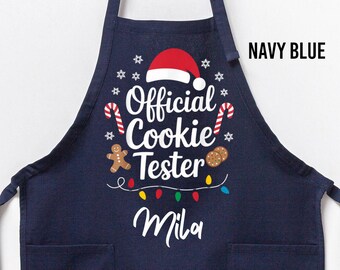 Op de afbeelding: Marineblauwe schort met de tekst "Official Cookie Tester" en de naam "Mila" in witte letters. De schort heeft kerstafbeeldingen, waaronder een kerstmuts, zuurstokken en een peperkoekmannetje. Het heeft twee voorzakken.