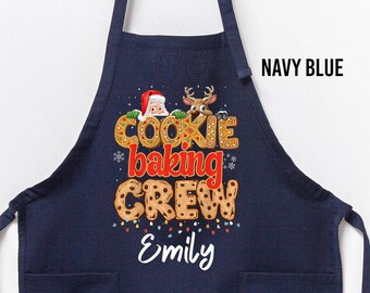 May include: Navy blue apron with two front pockets and a wooden hanger. The apron features the text "COOKIE BAKING CREW" in a festive font, with a Santa Claus and reindeer graphic. The name "Emily" is written below the text.