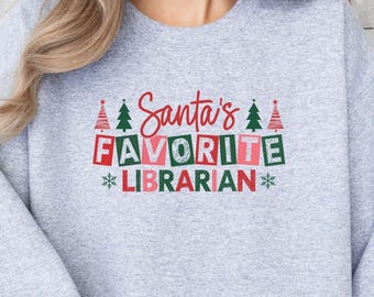 Puede incluir: Una sudadera gris claro con el texto "Santa's FAVORITE LIBRARIAN" en rojo, verde y blanco. El diseño incluye árboles de Navidad y copos de nieve. La sudadera es de cuello redondo.