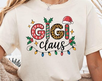 Puede incluir: Camiseta color crema con el texto "Gigi Claus" en un diseño festivo. Las letras están decoradas con patrones, acebo y luces navideñas. Un gorro de Papá Noel está sobre la letra "I".