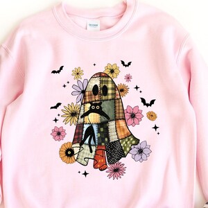 May include: Pink sweatshirt featuring a patchwork ghost design with floral and bat accents. The ghost has a sad expression. The word "PINK" is printed at the bottom. The sweatshirt is paired with striped flats.