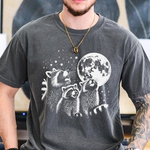 May include: A dark gray t-shirt with a graphic of three raccoons looking up at a full moon. The moon is white with a gray surface. The raccoons are white with black markings. The shirt has a distressed look.