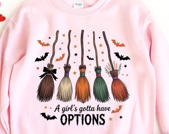 Op de afbeelding: Roze sweatshirt met een afbeelding van vijf heksenbezems in verschillende kleuren, met vleermuizen en sterren. De tekst "A girl's gotta have OPTIONS" staat eronder. Het woord "PINK" staat onderaan.