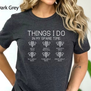 May include: A dark grey t-shirt with the text "THINGS I DO IN MY SPARE TIME" and a series of bee illustrations with text describing bee-related activities, such as "watch bees", "inspect beehives", "research bees", "talk about bees", "think about bees", and "dream about bees".