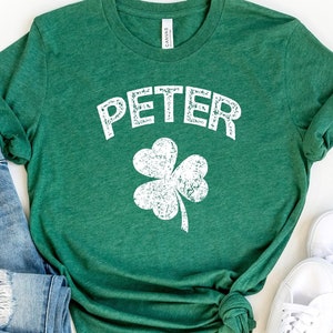 May include: Green t-shirt with a white shamrock and the word "PETER" in a distressed white font. The shirt is on a white background with a pair of white sneakers and blue jeans.