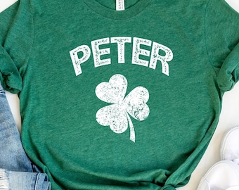 Op de afbeelding: Groen T-shirt met een wit klavertje vier en het woord "PETER" in een verweerd wit lettertype. Het shirt ligt op een witte achtergrond met een paar witte sneakers en een blauwe jeans.