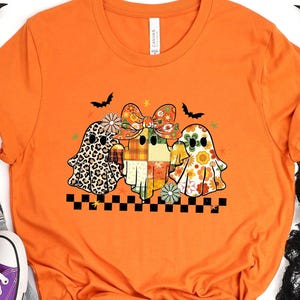 May include: Burnt orange t-shirt featuring a graphic of three ghost characters with floral, plaid, and leopard print designs. The shirt has a black and white checkered border at the bottom. The word "BURNT ORANGE" is printed at the top.