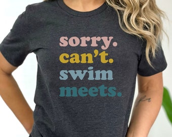 Puede incluir: Una camiseta gris oscuro con el texto "sorry. can't. swim meets." impreso en letras de colores.