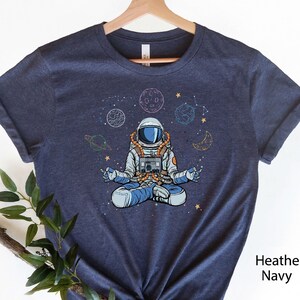 May include: A navy blue t-shirt with a graphic of an astronaut meditating in space. The astronaut is surrounded by planets, stars, and a crescent moon. The text "Heather Navy" is visible in the bottom right corner of the image.