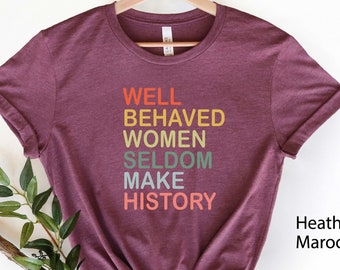 Op de afbeelding: Een maronkleurig T-shirt met de tekst "WELL BEHAVED WOMEN SELDOM MAKE HISTORY" gedrukt in regenboogkleuren.