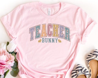 Op de afbeelding: Roze T-shirt met de tekst "TEACHER BUNNY" in regenboogkleuren. De tekst is in een retro-lettertype met konijnenoren aan beide kanten van het woord "BUNNY".