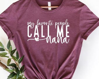 Puede incluir: Una camiseta granate con el texto blanco "my favorite people CALL ME Nana". La camiseta se muestra en una percha de madera con una ramita de vegetación en la esquina inferior izquierda. El texto incluye un símbolo de corazón.