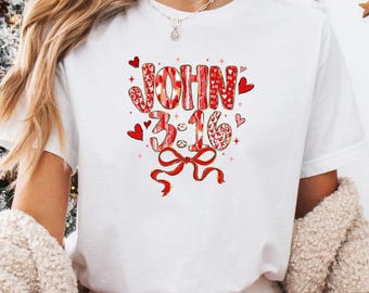 Puede incluir: Camiseta blanca con la inscripción "JOHN 3:16" en rojo y blanco, adornada con corazones y un lazo. El diseño forma parte de una temática navideña, ideal para la temporada festiva.