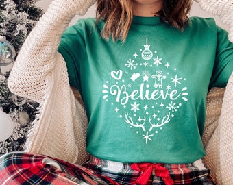 Op de afbeelding: Een vrouw draagt een groen T-shirt met een wit ontwerp met de tekst "Believe" omringd door kerstversiering en sneeuwvlokken. Ze draagt een rode en zwarte geruite pyjamabroek en een witte cardigan.