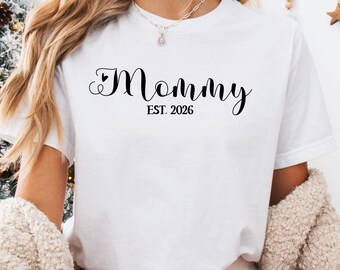 Op de afbeelding: Wit T-shirt met het woord "Mommy" in een scriptlettertype, met "EST. 2026" eronder. De persoon draagt een rood-witte geruite pyjamabroek en een crèmekleurige trui.