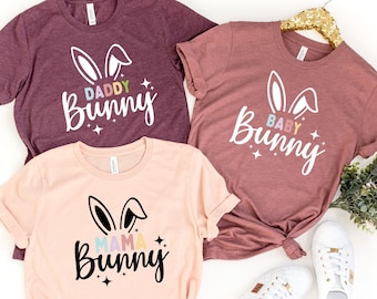 Op de afbeelding: Drie T-shirts met verschillende konijnenontwerpen. De shirts zijn maroon, roze en lichtroze. De ontwerpen zeggen "Daddy Bunny", "Baby Bunny" en "Mama Bunny".