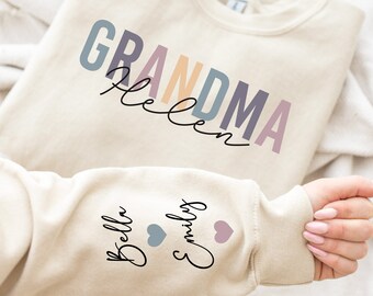 Op de afbeelding: Een beige sweatshirt met de tekst "Grandma Helen" in kleurrijke letters op de voorkant. De mouw heeft de namen "Bella" en "Emily" met hartjes.