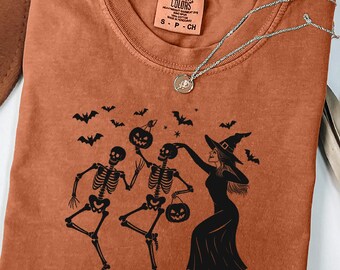 Op de afbeelding: Een oranje t-shirt met een Halloween-ontwerp. De afbeelding toont dansende skeletten, een heks, vleermuizen en pompoenen. Het shirt heeft een ronde hals en een klein label bij de halslijn. Een zilveren ketting is over het shirt gedrapeerd.