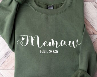 Op de afbeelding: Militairgroen sweatshirt met het woord "Memaw" in een sierlijk lettertype, een hartje en de tekst "EST. 2026". Het sweatshirt is opgevouwen, met de mouwen zichtbaar. De tekst "MILITARY GREEN" staat onderaan.