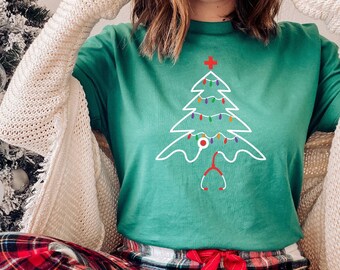 Op de afbeelding: Groen t-shirt met een kerstboomontwerp, met een kruis, lichtjes en een stethoscoop. De persoon draagt een rood-zwarte geruite pyjamabroek. Een wit vest is over de schouders gedrapeerd. Een kerstboom met witte ballen op de achtergrond.
