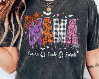 Op de afbeelding: Een antracietgrijs T-shirt met korte mouwen en het woord MAMA in grote, gedessineerde letters. De M is paars met een strik, de A is oranje met pompoenen, de M is zwart-wit geblokt en de A is roze met luipaardprint. Daaronder staan de namen Emma, Noah en Sarah met spookpictogrammen.