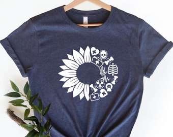 Puede incluir: Una camiseta azul marino jaspeada con un diseño de girasol blanco. El girasol está formado por símbolos médicos, incluyendo un corazón, una cruz, una calavera y huesos cruzados, una mano, una caja torácica, un latido del corazón, un símbolo de radiación y un botiquín de primeros auxilios.