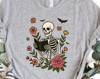 Op de afbeelding: Heather athletic grey t-shirt met een skelet dat een boek leest, omringd door bloemen, bladeren en vleermuizen. De tekst "HEATHER ATHLETIC GREY" staat onderaan de shirt. Een casual, comfortabel ontwerp.