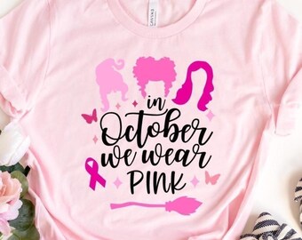Op de afbeelding: Lichtroze T-shirt met de tekst "in October we wear PINK" en een afbeelding van drie heksensilhouetten, een roze lint, vlinders en een bezem. Het woord "PINK" staat onderaan gedrukt. Gestreepte ballerina's liggen ernaast.
