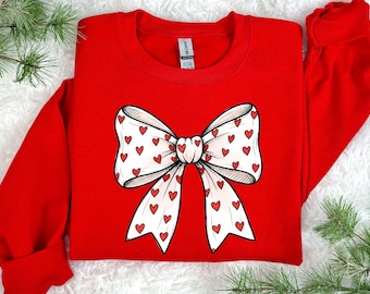 Puede incluir: Una sudadera roja con un gran lazo blanco con patrones de corazones rojos. El lazo es un elemento de diseño central, con líneas detalladas. La sudadera es de color rojo liso, con mangas largas y cuello redondo.