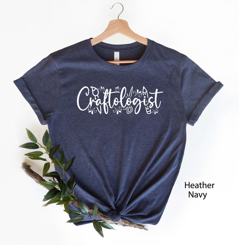 May include: A heather navy blue t-shirt with the text "Craftologist" in white lettering. The text is surrounded by small white floral and craft-related designs.